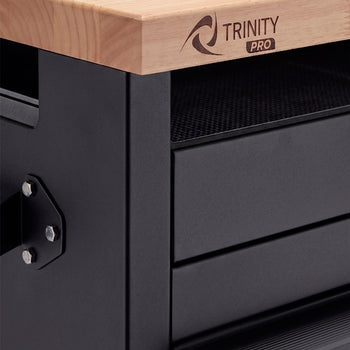 A close-up shot highlights the front-left corner of a matte black metal workbench frame beneath a solid wood countertop. The front corner of the butcher block surface is laser-engraved with the "TRINITY PRO" logo.