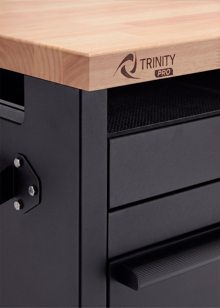 A close-up shot highlights the front-left corner of a matte black metal workbench frame beneath a solid wood countertop. The front corner of the butcher block surface is laser-engraved with the "TRINITY PRO" logo.