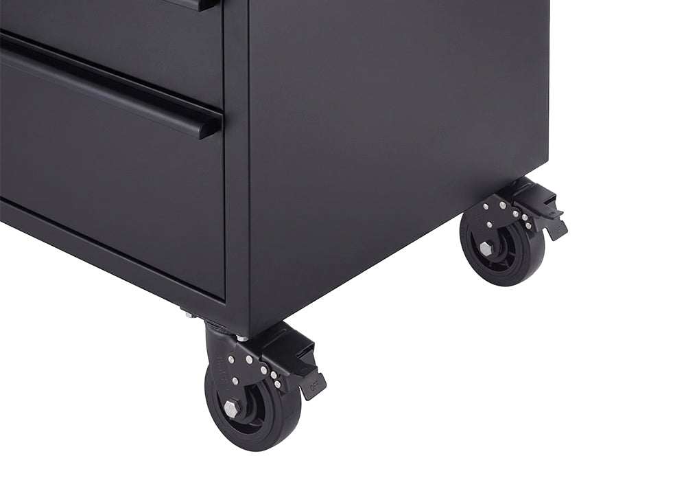 A close-up shot focuses on the lower corner of a matte black tool workbench showing its heavy-duty caster wheels. The dark swivel wheels feature integrated foot-operated brake levers to lock the unit securely in place.