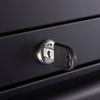 A close-up view focuses on a locking mechanism integrated into the matte black metal panel of a workbench. A key with a black molded plastic head is inserted into the cylinder lock to secure the drawers.