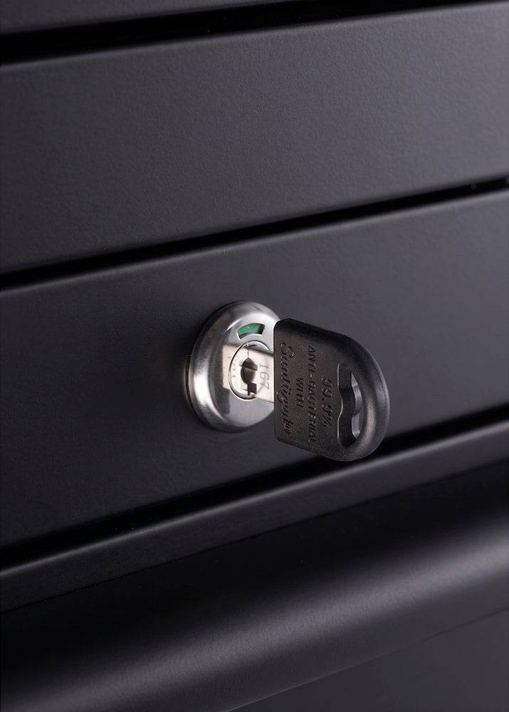 A close-up view focuses on a locking mechanism integrated into the matte black metal panel of a workbench. A key with a black molded plastic head is inserted into the cylinder lock to secure the drawers.
