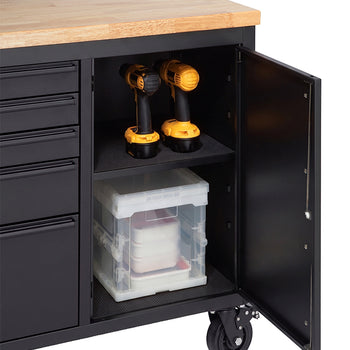 A close-up shot highlights the open side cabinet of a matte black mobile workbench with a solid wooden top. Two cordless drills sit on the lined upper shelf, while a clear plastic storage container is organized on the lower shelf.
