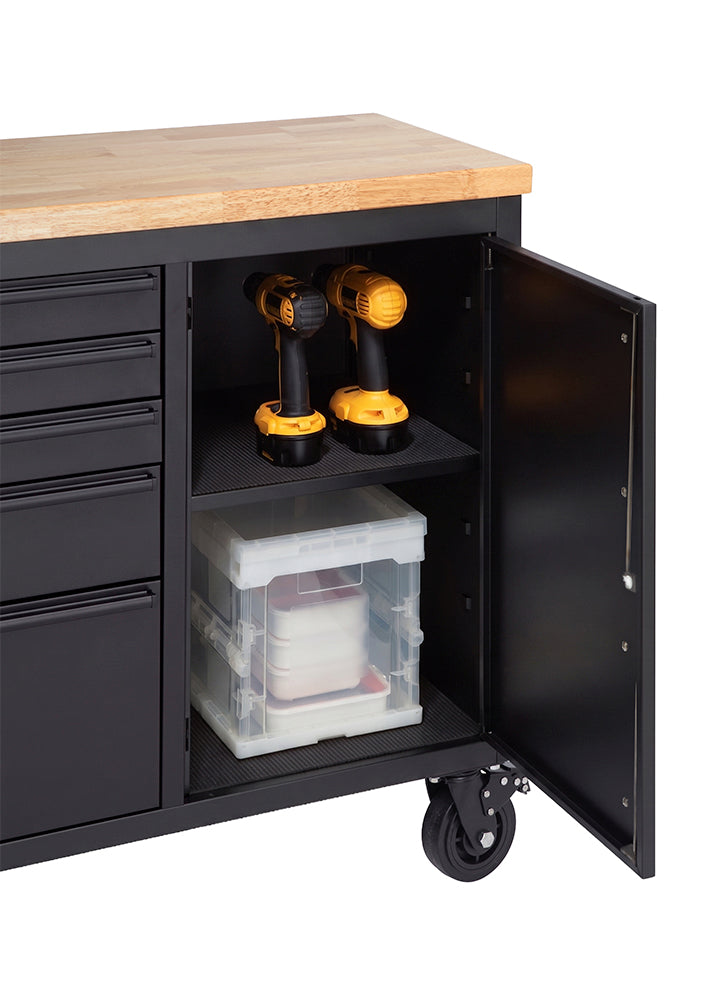 A close-up shot highlights the open side cabinet of a matte black mobile workbench with a solid wooden top. Two cordless drills sit on the lined upper shelf, while a clear plastic storage container is organized on the lower shelf.