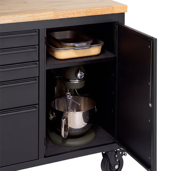 A close-up shot highlights the open side cabinet of a matte black workbench with a solid wood countertop. The interior shelf holds stacked baking pans on the top tier, while a large stand mixer sits securely on the bottom tier.