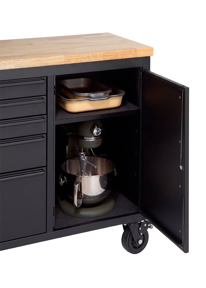 A close-up shot highlights the open side cabinet of a matte black workbench with a solid wood countertop. The interior shelf holds stacked baking pans on the top tier, while a large stand mixer sits securely on the bottom tier.