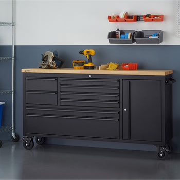 A matte black mobile tool workbench with a light wood top is set against a two-tone garage wall with all drawers and cabinets securely closed. Power tools like a circular saw and cordless drill rest on the surface, flanked by wire storage racks and wall-mounted bin organizers.