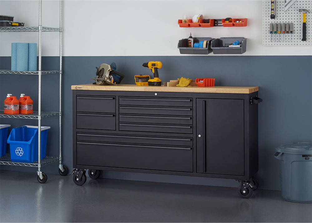 A matte black mobile tool workbench with a light wood top is set against a two-tone garage wall with all drawers and cabinets securely closed. Power tools like a circular saw and cordless drill rest on the surface, flanked by wire storage racks and wall-mounted bin organizers.