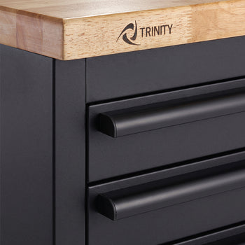 A close-up shot shows the upper-left corner of a matte black tool workbench featuring stacked drawer handles. A smooth wooden countertop rests above the drawers with the "TRINITY" logo branded onto its wooden edge.