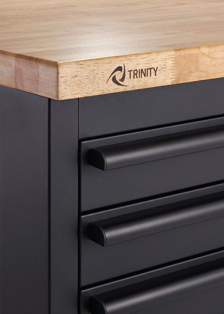 A close-up shot shows the upper-left corner of a matte black tool workbench featuring stacked drawer handles. A smooth wooden countertop rests above the drawers with the "TRINITY" logo branded onto its wooden edge.