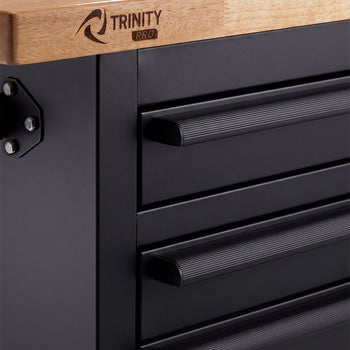 A close-up shot highlights the corner of a black tool cabinet featuring a thick wooden workbench top with a branded "TRINITY PRO" logo. The matte black drawers are equipped with textured, ridged horizontal pull handles for an easy grip.