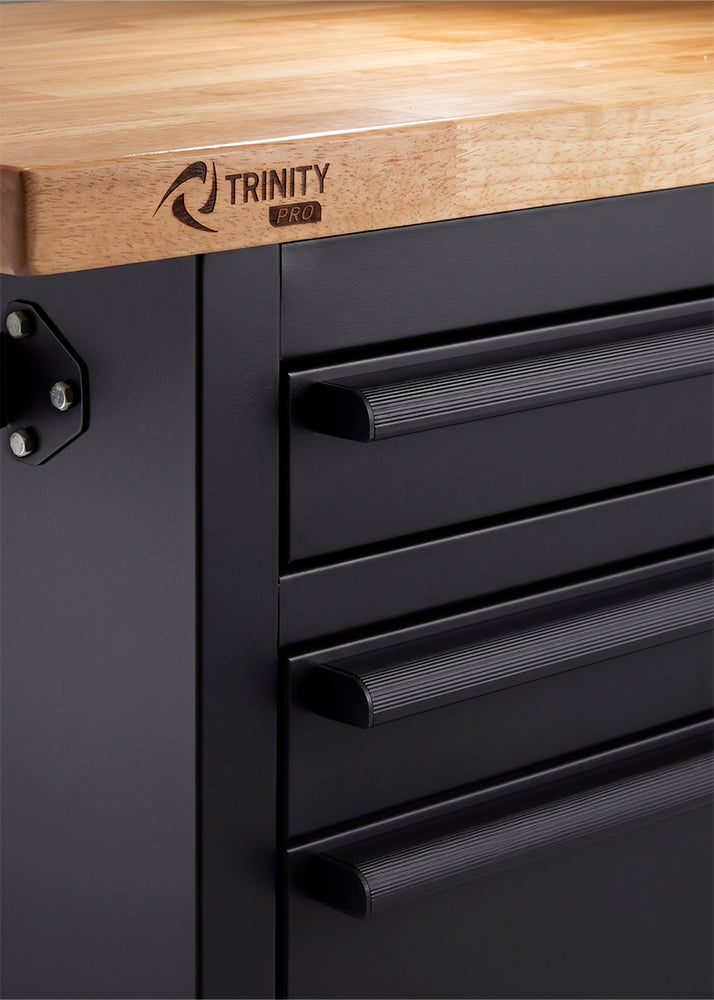 A close-up shot highlights the corner of a black tool cabinet featuring a thick wooden workbench top with a branded "TRINITY PRO" logo. The matte black drawers are equipped with textured, ridged horizontal pull handles for an easy grip.