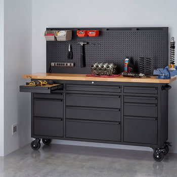 A black rolling workbench with a wooden top and integrated pegboard is organized with tools, small storage bins, and a blue vise in a workshop setting. One of the left drawers is pulled open to reveal a cordless drill, while a red air compressor sits on the floor nearby.