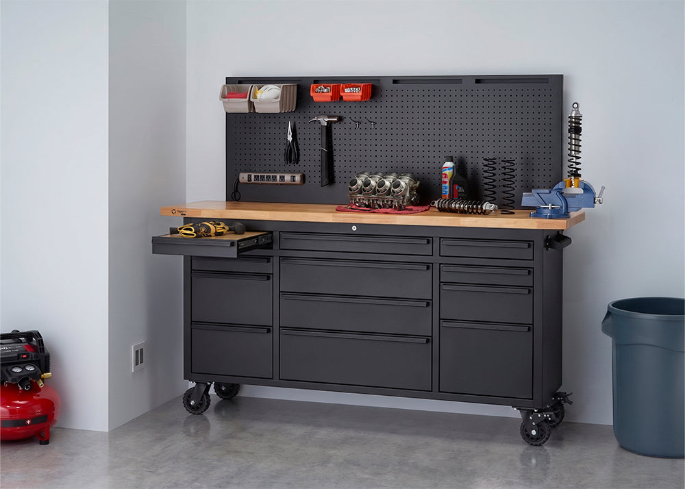 A black rolling workbench with a wooden top and integrated pegboard is organized with tools, small storage bins, and a blue vise in a workshop setting. One of the left drawers is pulled open to reveal a cordless drill, while a red air compressor sits on the floor nearby.