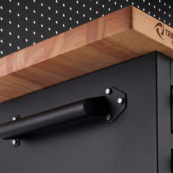 A close-up view highlights a sturdy, matte black handle bolted to the side of a black tool cabinet. Just above the handle, the edge of a thick wooden workbench top features the branded "TRINITY PRO" logo.