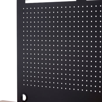A close-up shot highlights a black metal pegboard with a grid of small holes, mounted to a light wood workbench. The panel features wide horizontal cutouts along the top edge and a circular grommet at the bottom corner for clean cable management.