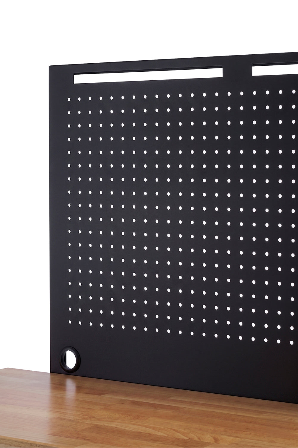 A close-up shot highlights a black metal pegboard with a grid of small holes, mounted to a light wood workbench. The panel features wide horizontal cutouts along the top edge and a circular grommet at the bottom corner for clean cable management.