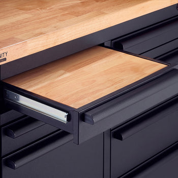 A close-up view of a black Trinity Pro workbench shows a shallow drawer pulled open to reveal a light wood inlay. The drawer slides on metal glides just below the matching solid wood top and is fitted with a textured black pull handle.