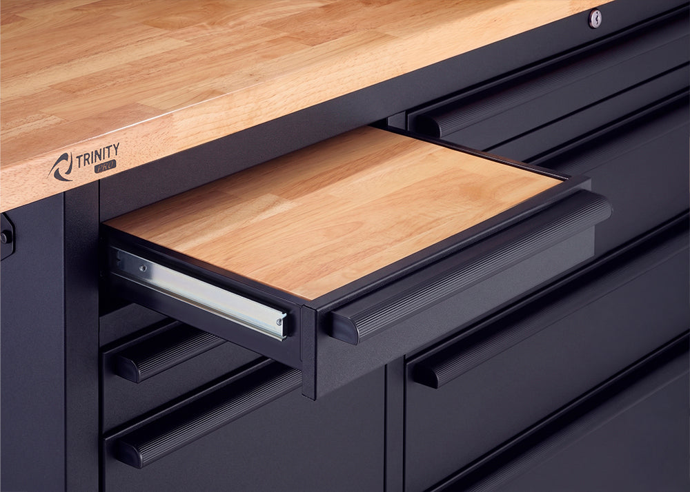 A close-up view of a black Trinity Pro workbench shows a shallow drawer pulled open to reveal a light wood inlay. The drawer slides on metal glides just below the matching solid wood top and is fitted with a textured black pull handle.