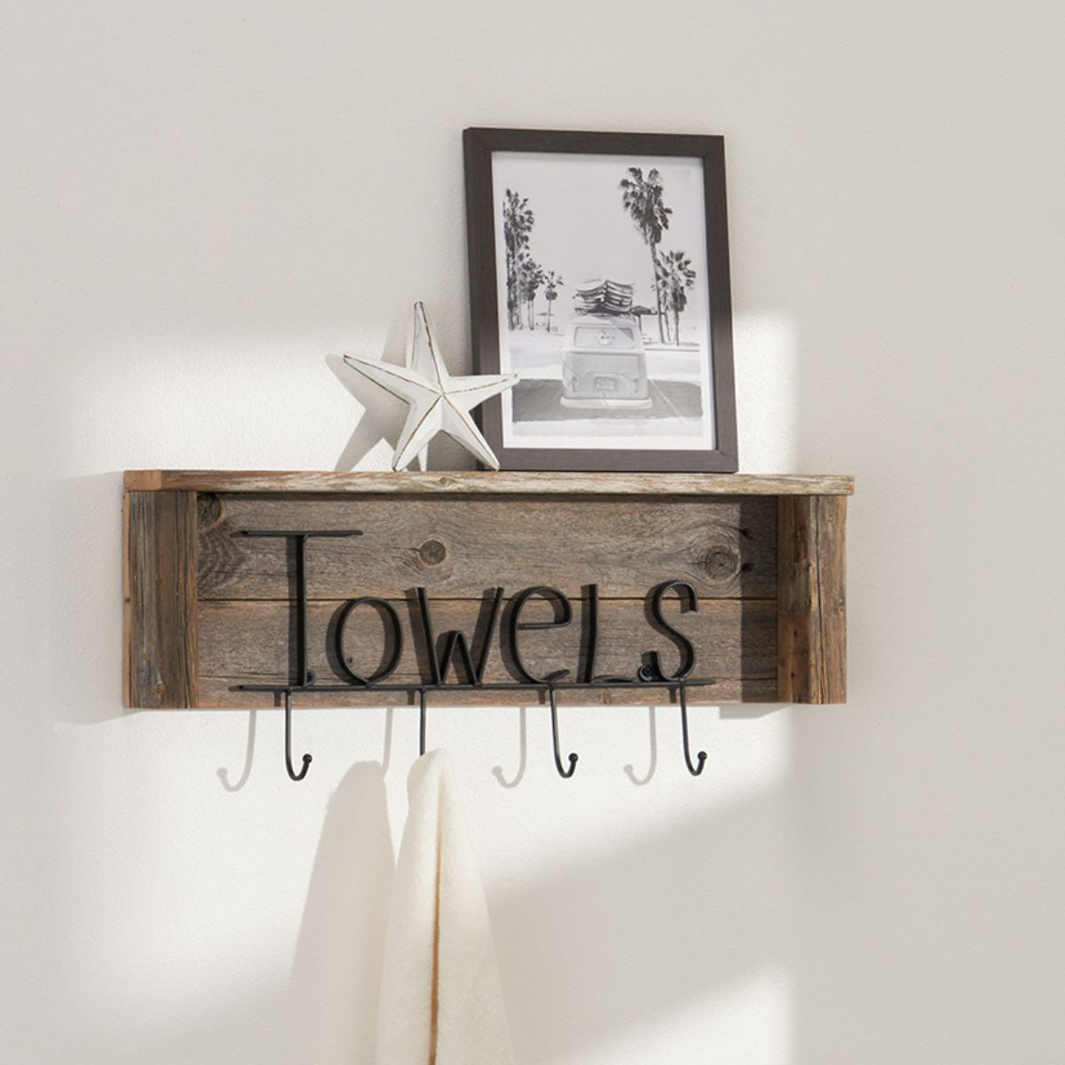 A rustic wooden shelf with black metal lettering spelling "Towels" and five hanging hooks is mounted on a light beige wall. Above the shelf, a white starfish and a framed black and white photograph of palm trees and a vehicle rest on the shelf's surface.