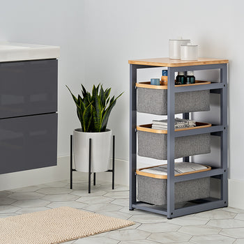 A three-tier gray storage unit with a bamboo top and fabric baskets sits next to a potted snake plant in a modern bathroom. The room features white walls, hexagonal floor tiles, and a sleek charcoal-colored vanity.