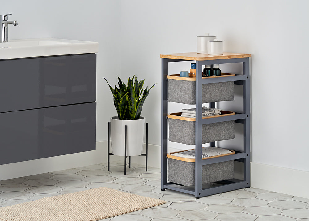 A three-tier gray storage unit with a bamboo top and fabric baskets sits next to a potted snake plant in a modern bathroom. The room features white walls, hexagonal floor tiles, and a sleek charcoal-colored vanity.