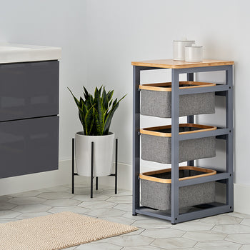 A modern bathroom features a sleek gray vanity and a three-tier storage unit with a bamboo top and gray fabric baskets. A potted snake plant stands between the furniture on a white hexagonal tile floor, adding a touch of greenery to the clean, minimalist space.