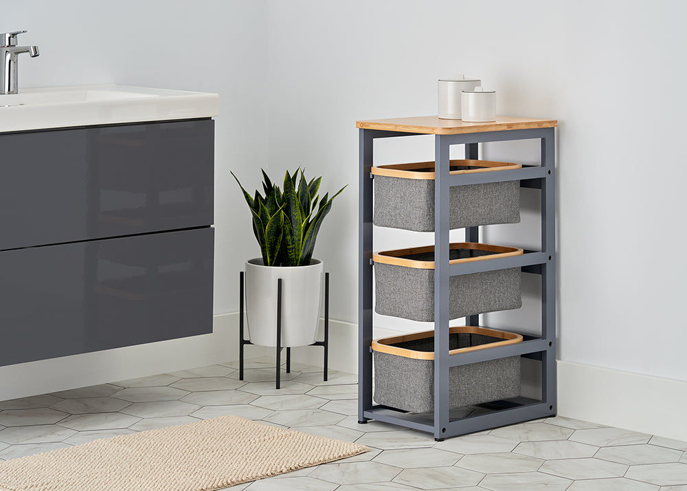 A modern bathroom features a sleek gray vanity and a three-tier storage unit with a bamboo top and gray fabric baskets. A potted snake plant stands between the furniture on a white hexagonal tile floor, adding a touch of greenery to the clean, minimalist space.