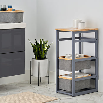 A modern bathroom corner features a dark gray floating vanity and a matching gray tiered storage rack with a light wood top and fabric baskets. A potted snake plant sits between them on a black metal stand, set against light gray walls and hexagonal floor tiles.