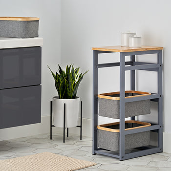 A modern bathroom corner features a grey vanity and a matching slim storage tower with two fabric baskets and a wooden top. A potted snake plant stands in the corner against a white wall, complemented by light grey hexagonal floor tiles and a neutral woven rug.