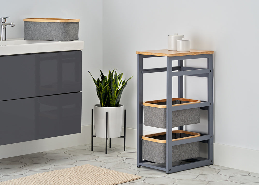A modern bathroom corner features a grey vanity and a matching slim storage tower with two fabric baskets and a wooden top. A potted snake plant stands in the corner against a white wall, complemented by light grey hexagonal floor tiles and a neutral woven rug.