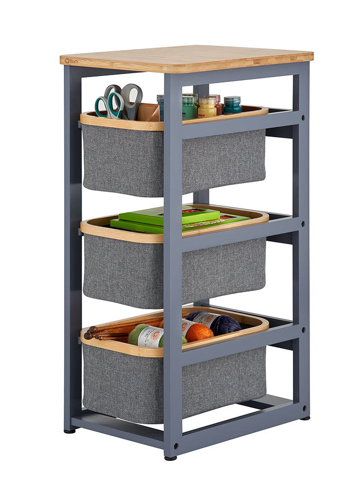 A three-tier storage organizer features a grey metal frame with a light wood top and three removable grey fabric baskets rimmed in wood. Each basket is filled with various craft supplies, including scissors, paint jars, books, and colorful yarn.