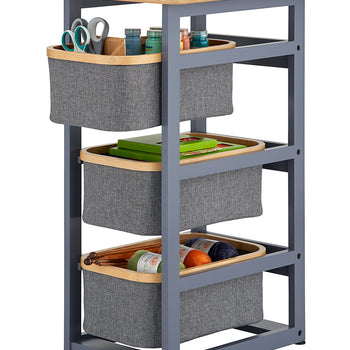 A three-tier craft storage cart features a grey metal frame with a light wood top and three slide-out grey fabric bins with wooden rims. Each bin is filled with various hobby supplies, including scissors, paint bottles, green boxes, and balls of colorful yarn.