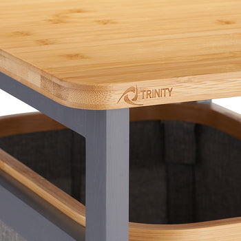 A close-up shot shows a light-colored bamboo tabletop with a rounded corner featuring a laser-etched "TRINITY" logo. Below the surface, a dark gray metal frame supports a sliding fabric laundry hamper with a matching wood-trimmed rim.