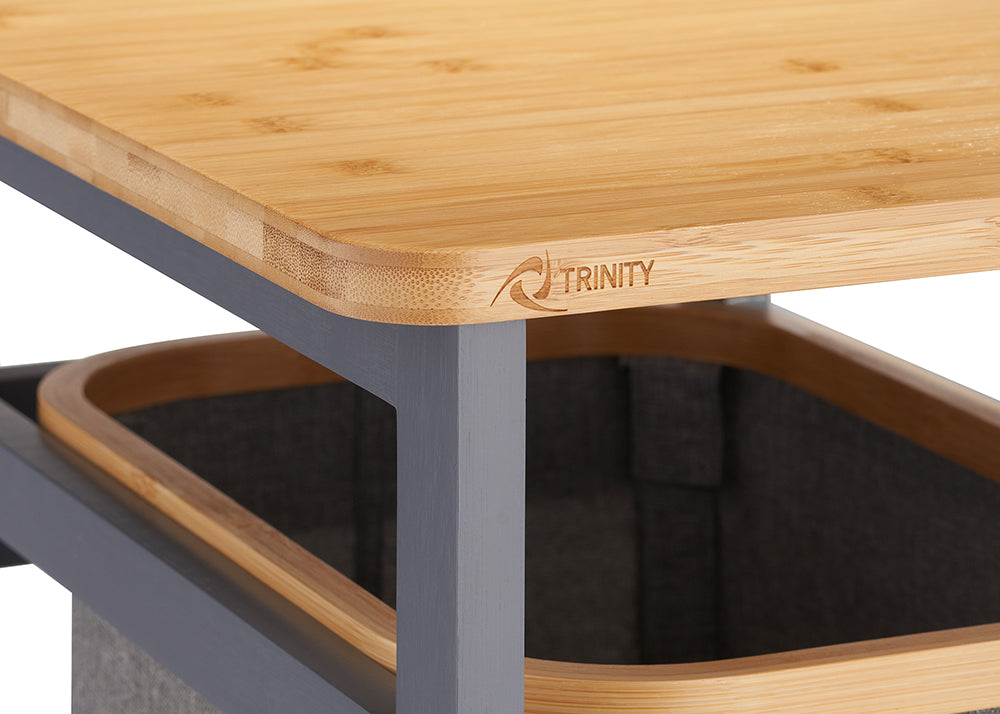 A close-up shot shows a light-colored bamboo tabletop with a rounded corner featuring a laser-etched "TRINITY" logo. Below the surface, a dark gray metal frame supports a sliding fabric laundry hamper with a matching wood-trimmed rim.