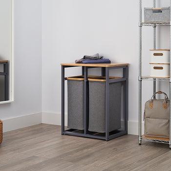 A stylish laundry sorter with two grey fabric bags and a wooden top sits on a wood-style floor. A tall chrome shelf unit with storage boxes and hanging clothes is next to it, and a large mirror leans against the opposite wall.