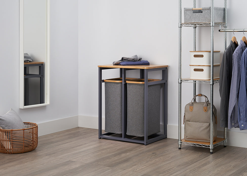 A stylish laundry sorter with two grey fabric bags and a wooden top sits on a wood-style floor. A tall chrome shelf unit with storage boxes and hanging clothes is next to it, and a large mirror leans against the opposite wall.