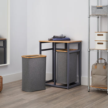 A clean, modern laundry room is shown with a chrome-wire shelving unit and a mirror. In the center, there is a pair of gray fabric laundry hampers, one with a lid and one on a metal frame with a bamboo top.