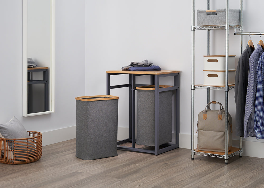 A clean, modern laundry room is shown with a chrome-wire shelving unit and a mirror. In the center, there is a pair of gray fabric laundry hampers, one with a lid and one on a metal frame with a bamboo top.