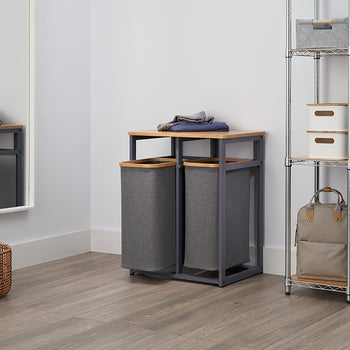 A clean, modern laundry room is shown with a chrome-wire shelving unit and a mirror. In the center, there is a pair of gray fabric laundry hampers, one with a lid and one on a metal frame with a bamboo top.