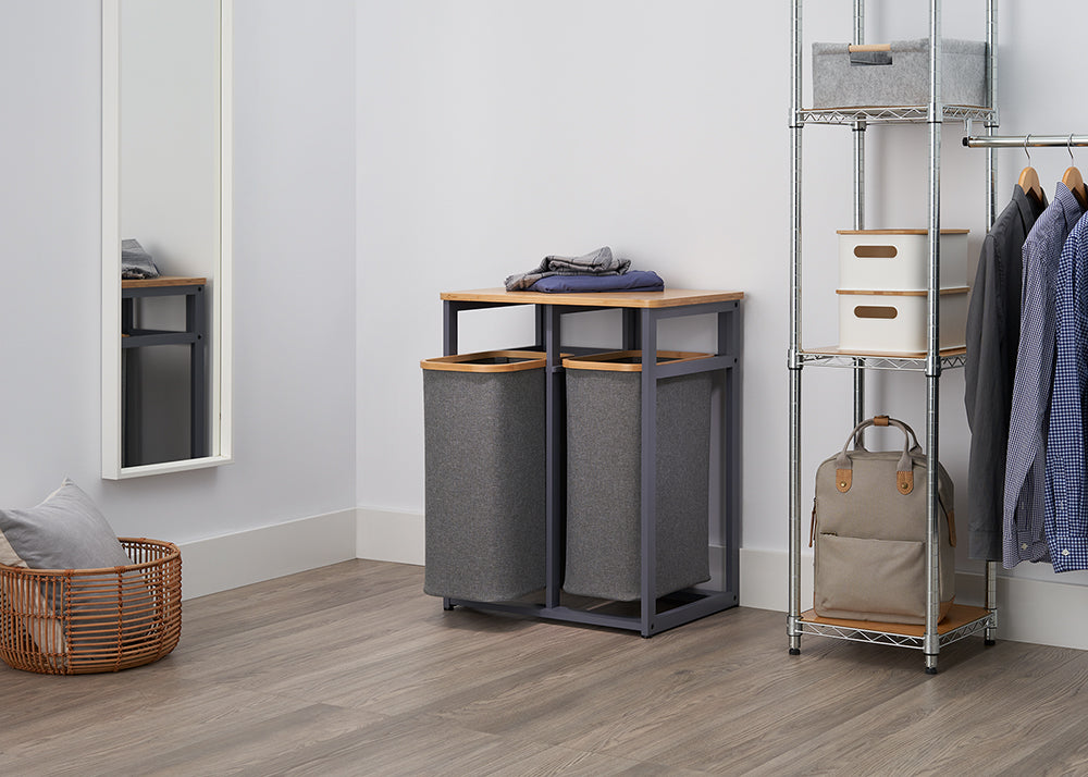 A clean, modern laundry room is shown with a chrome-wire shelving unit and a mirror. In the center, there is a pair of gray fabric laundry hampers, one with a lid and one on a metal frame with a bamboo top.