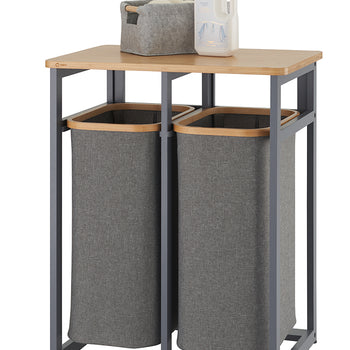 A laundry sorter with two grey fabric hampers is shown, suspended within a matte grey metal frame. A light-colored wood shelf sits on top, holding a small grey felt basket and a bottle of laundry detergent.