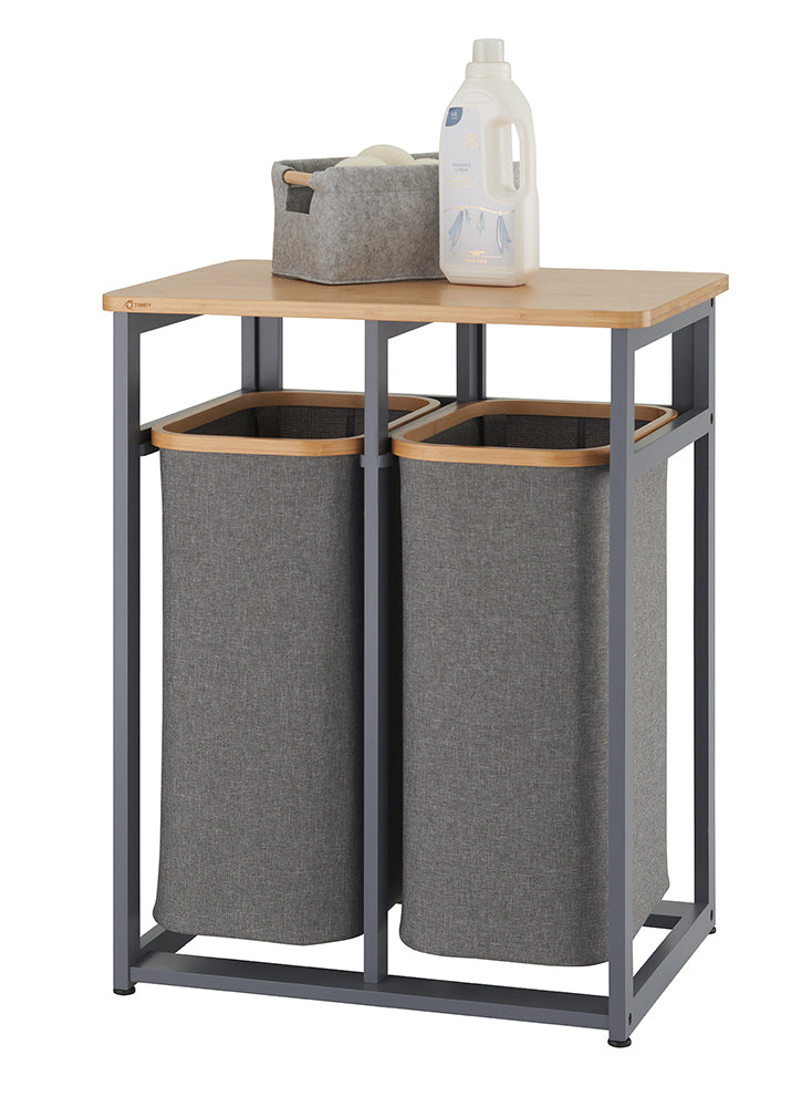 A laundry sorter with two grey fabric hampers is shown, suspended within a matte grey metal frame. A light-colored wood shelf sits on top, holding a small grey felt basket and a bottle of laundry detergent.