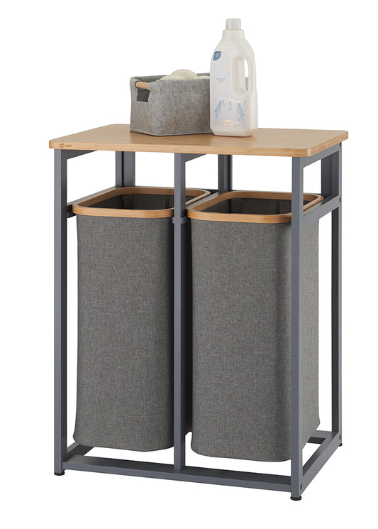 A laundry sorter with two grey fabric hampers is shown, suspended within a matte grey metal frame. A light-colored wood shelf sits on top, holding a small grey felt basket and a bottle of laundry detergent.