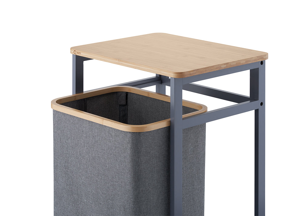 A close-up view demonstrates the gray fabric hamper bag pulled forward out of its dark gray metal frame, showcasing its wooden top rim. Above the hamper, a smooth wooden tabletop rests on the frame against a clean white background.