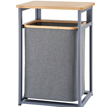 A standalone laundry hamper unit features a dark gray metal frame and a smooth wooden tabletop. Suspended within the frame is a removable gray fabric hamper bag with a wooden trim around the top rim.