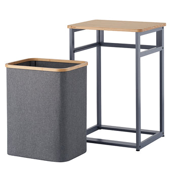 A gray fabric laundry hamper with light wood trim stands detached beside its dark gray metal frame. The frame features an open base design topped with a matching wooden tabletop on a plain white background.