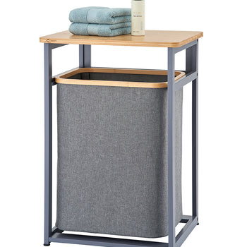 A freestanding laundry hamper unit features a dark gray metal frame and a light wooden tabletop with a removable gray fabric hamper bag below. Staked on top of the wooden shelf are two folded light blue towels alongside a pump bottle.