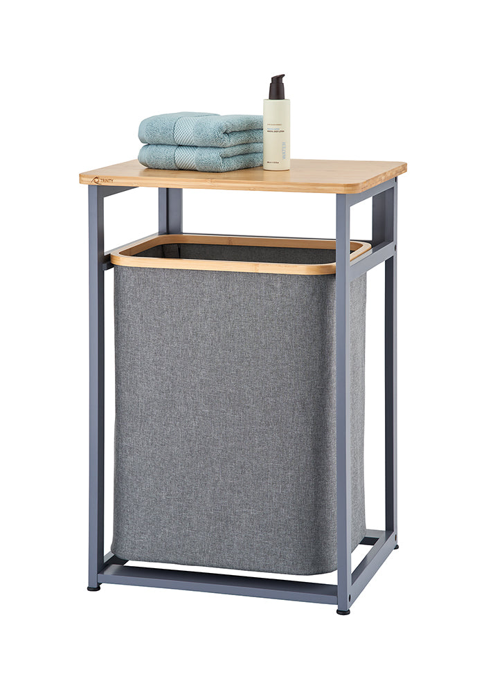 A freestanding laundry hamper unit features a dark gray metal frame and a light wooden tabletop with a removable gray fabric hamper bag below. Staked on top of the wooden shelf are two folded light blue towels alongside a pump bottle.