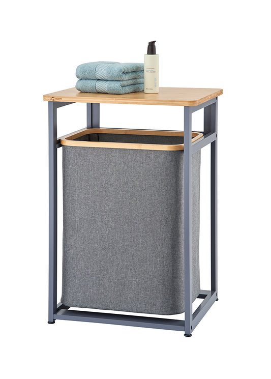 A freestanding laundry hamper unit features a dark gray metal frame and a light wooden tabletop with a removable gray fabric hamper bag below. Staked on top of the wooden shelf are two folded light blue towels alongside a pump bottle.