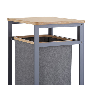 A close-up shot highlights the upper portion of a laundry hamper unit, focusing on its light wooden tabletop and dark gray metal frame. Directly beneath the top shelf, a gray fabric hamper bag with a wooden-trimmed rim rests suspended inside the structure.